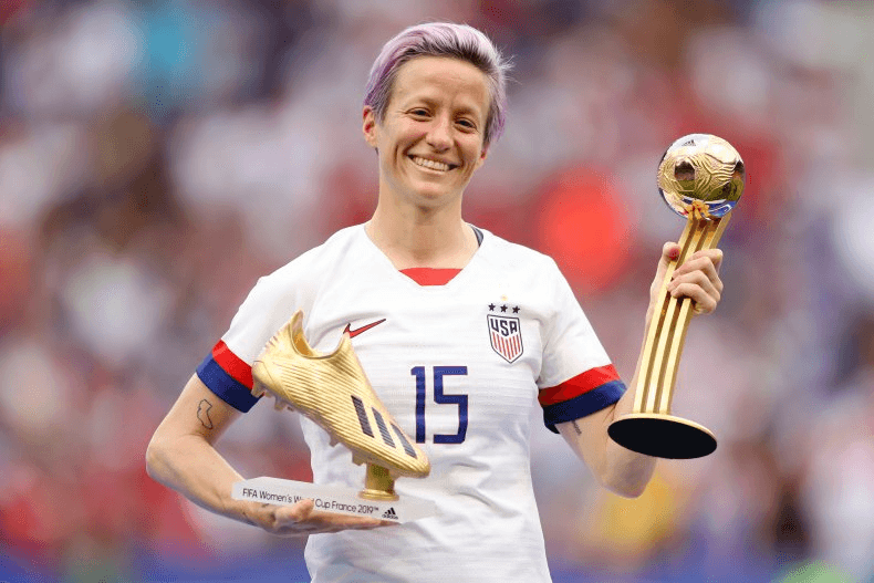 Megan Rapinoe holding 2 trophies on the field