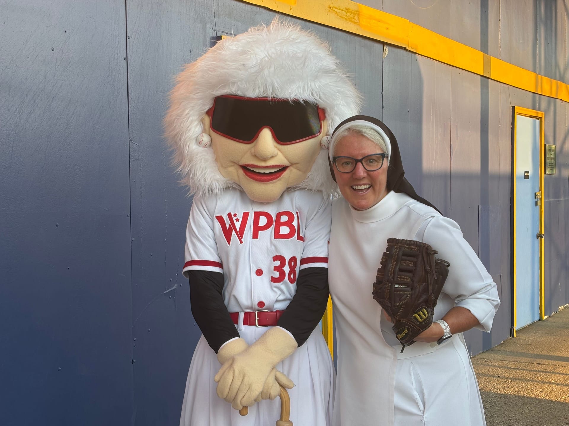 A smiling woman dressed in a white baseball-themed nun costume poses with a glove beside a WPBL mascot wearing a white uniform, oversized sunglasses, and a furry white headpiece.