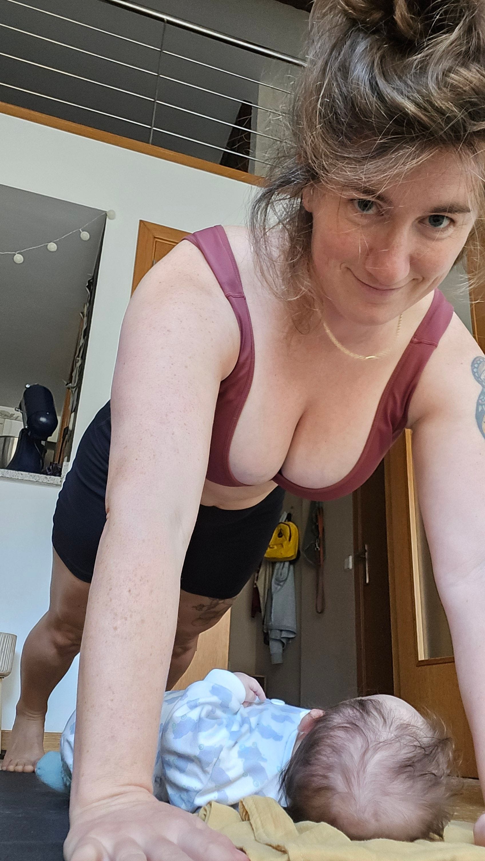 A woman in workout clothes holds a high plank position on a yoga mat while smiling toward the camera, with a baby lying on a blanket beneath her. The photo is taken indoors in a loft-style apartment with natural light, showing a candid postpartum workout and parenting moment.