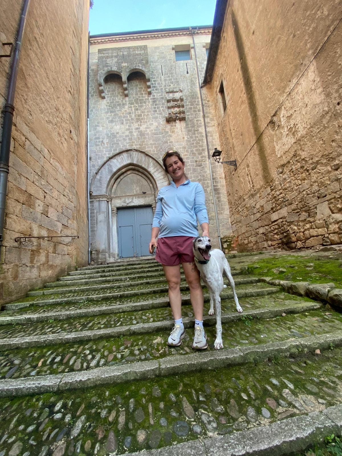 Smiling pregnant woman standing on mossy stone steps in a narrow historic alley, holding a leash attached to a white dog, with an old stone building and arched doorway behind her.