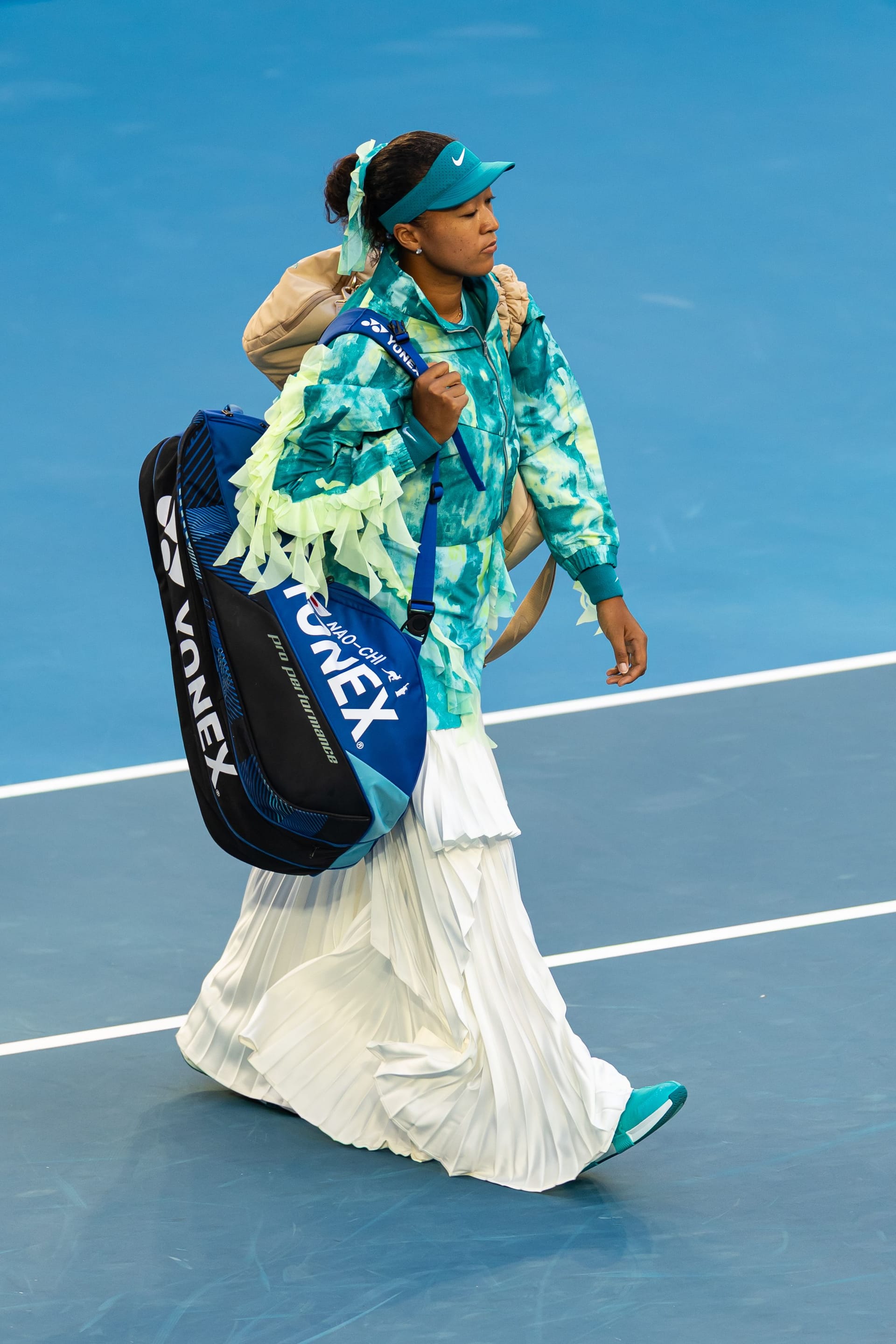 Naomi Osaka walks across a blue court carrying a Yonex racket bag while wearing an elaborate teal-and-white outfit with a pleated floor-length skirt, ruffled jacket, and matching visor.
