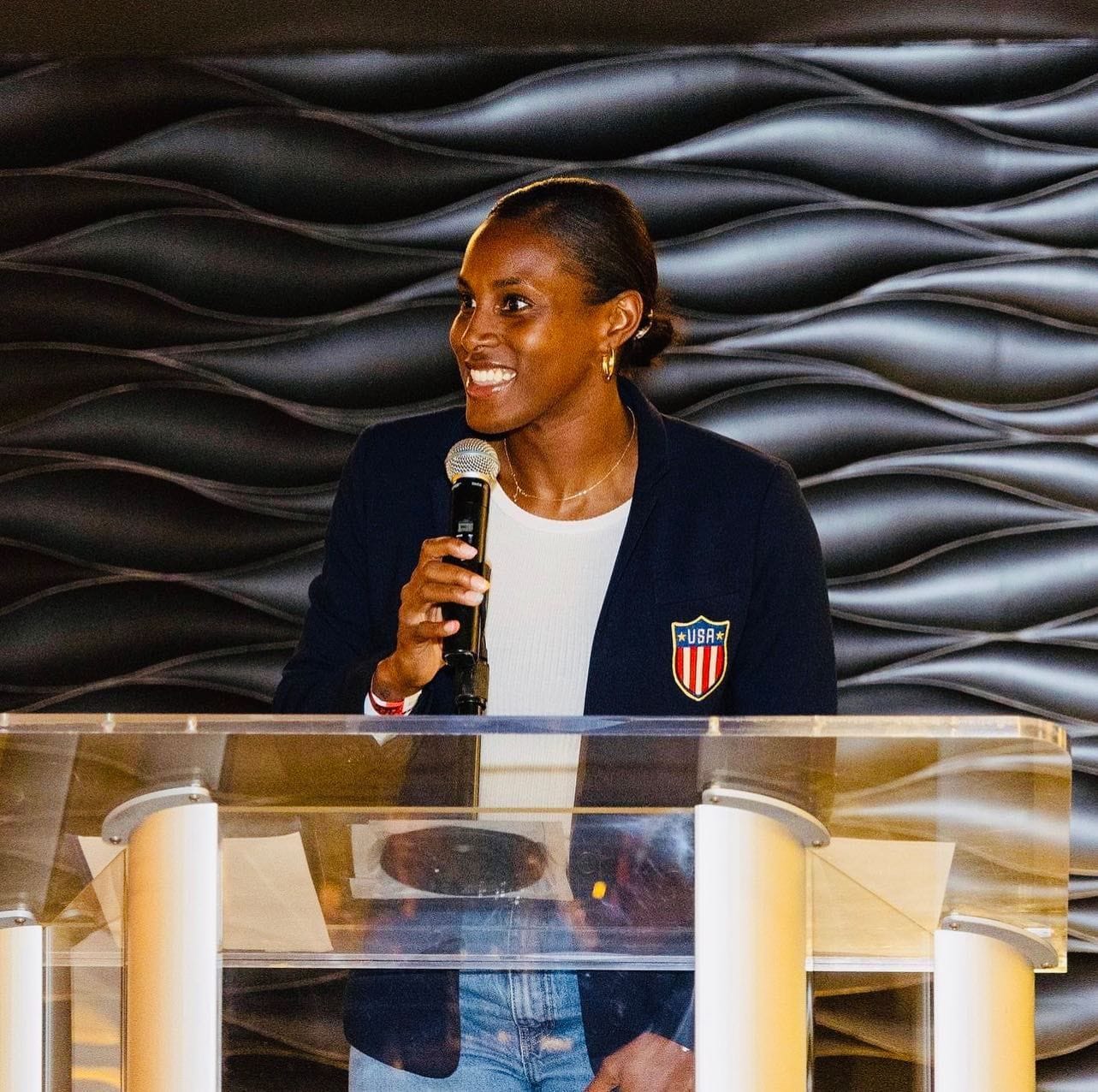 Naya speaks into a microphone at the podium for a Team USA event