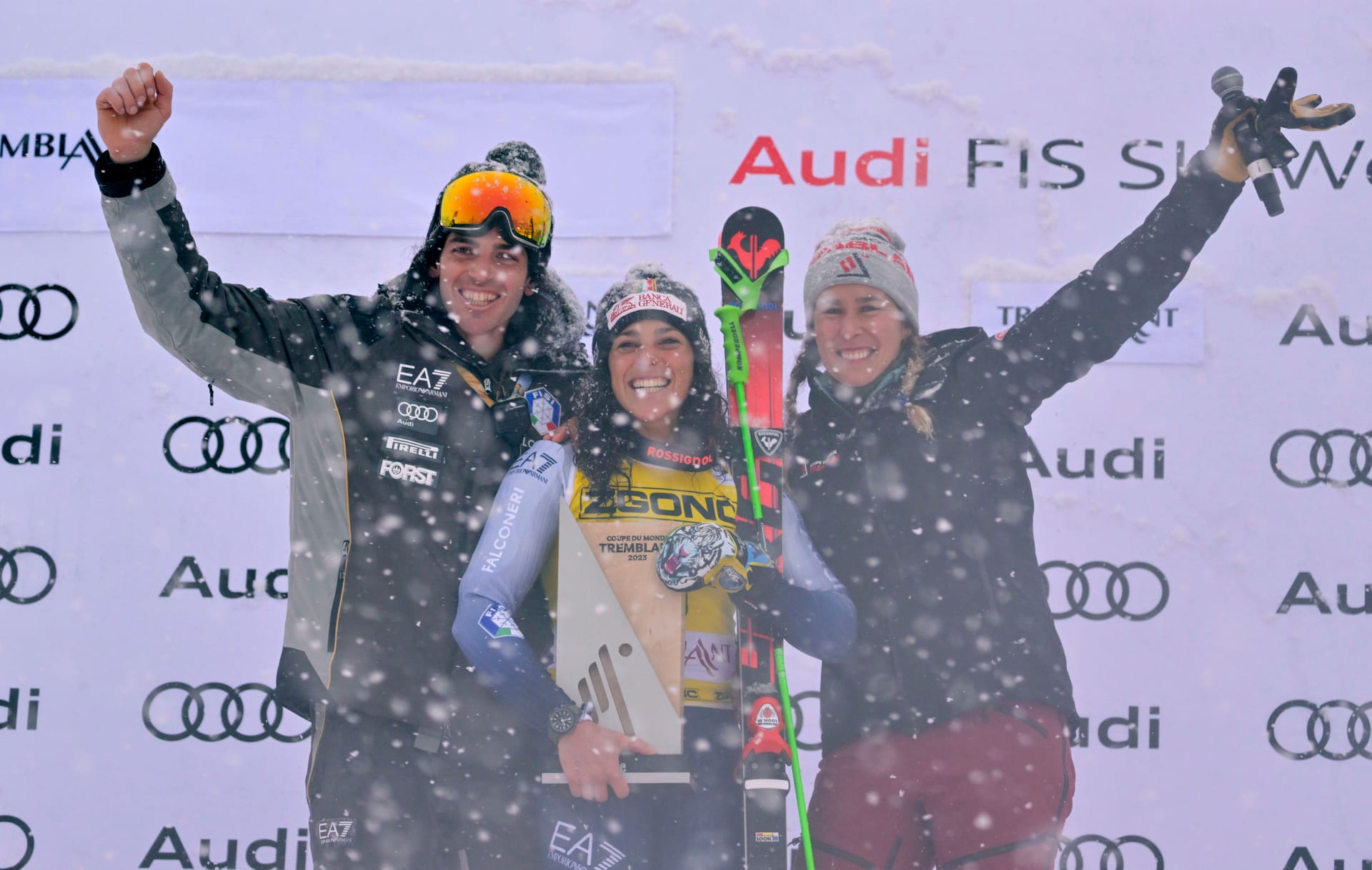 Female skiier smiles with a trophy on an event stage with 2 presenters