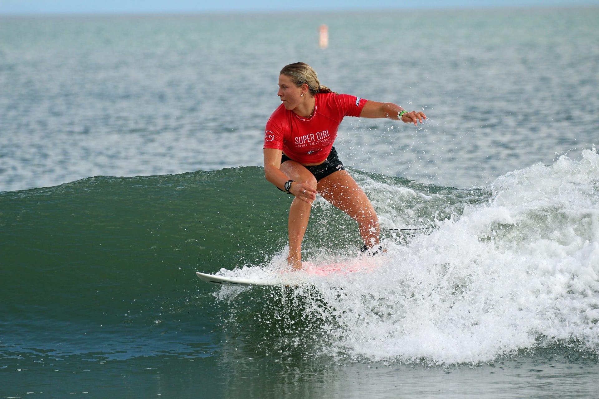 A surfer in a red competition jersey rides along the face of a breaking wave, crouched low on the board with water spraying around them. The ocean stretches into the background as the athlete maintains balance and speed during a surfing heat.